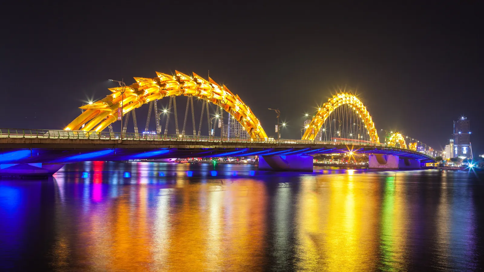 Dragon Bridge in Da Nang lit in gold and blue at night, with colorful reflections on the Han River