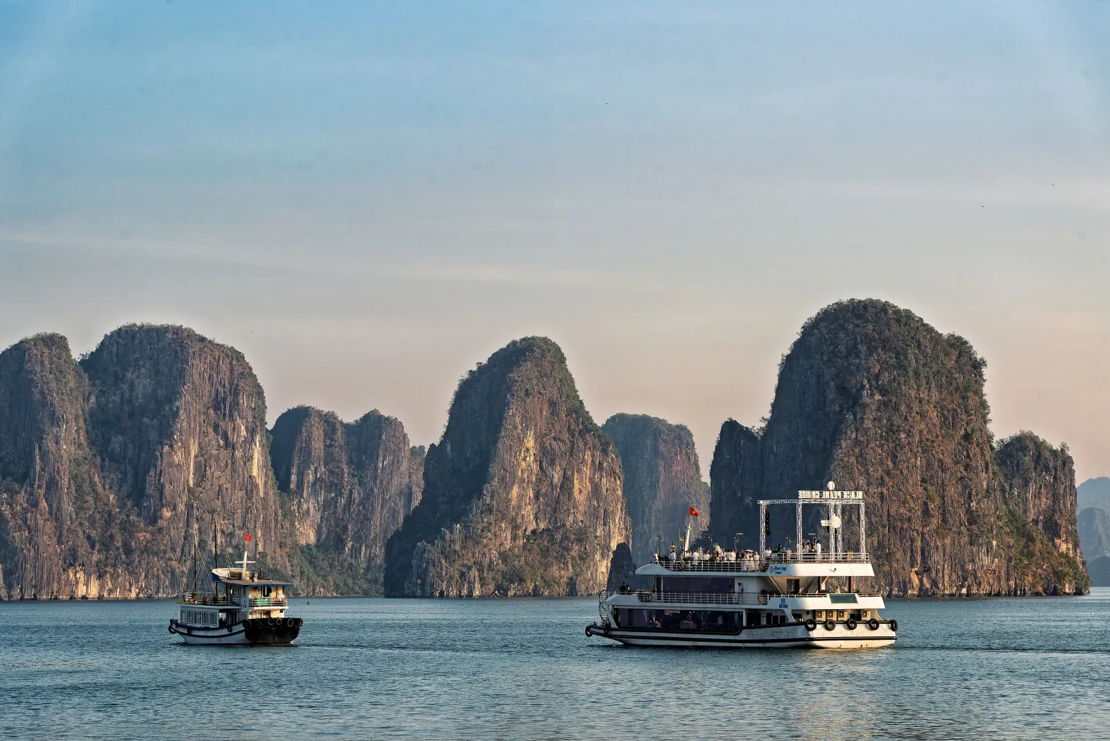 Limestone karst towers over calm water in Ha Long Bay with a small tour boat and a two-deck cruise boat in soft afternoon light