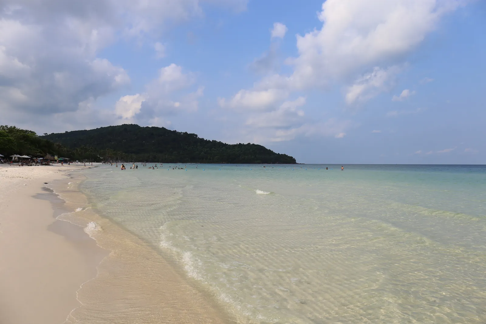富國島星沙灘的海岸線,左側是白色沙灘,右側是清澈的淺綠色海水,遠處有林木覆蓋的岬角與幾位戲水的人,天空有雲